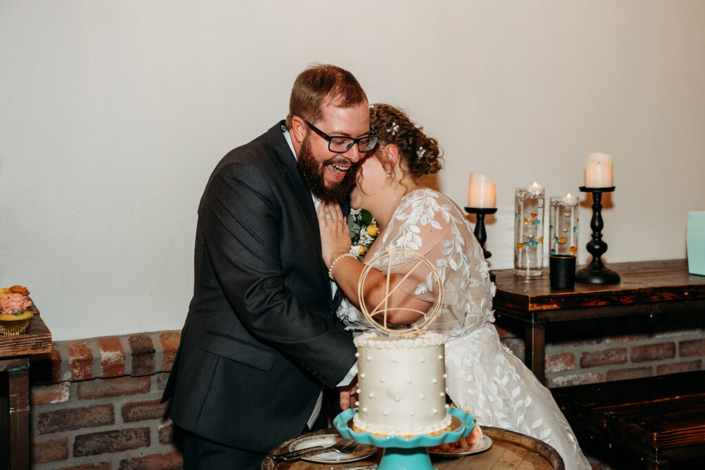 Bride and groom kissing before cutting the cake, cake is one tier with pearl dots with a simple wooden initial cake toppper