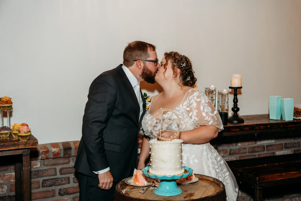 Bride and groom kissing before cutting the cake, cake is one tier with pearl dots with a simple wooden initial cake toppper