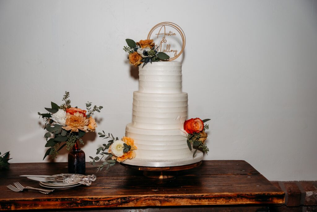 3 tiered white wedding cake with orange roses, dahlias and a gold name cake topper,