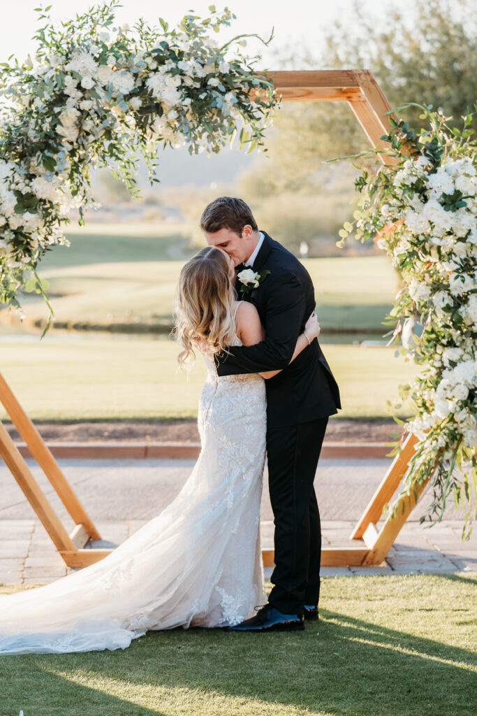 Groom kissing the bride at verrado golf club wedding ceremony, arizona wedding photographer, hexagon wood arch with white florals